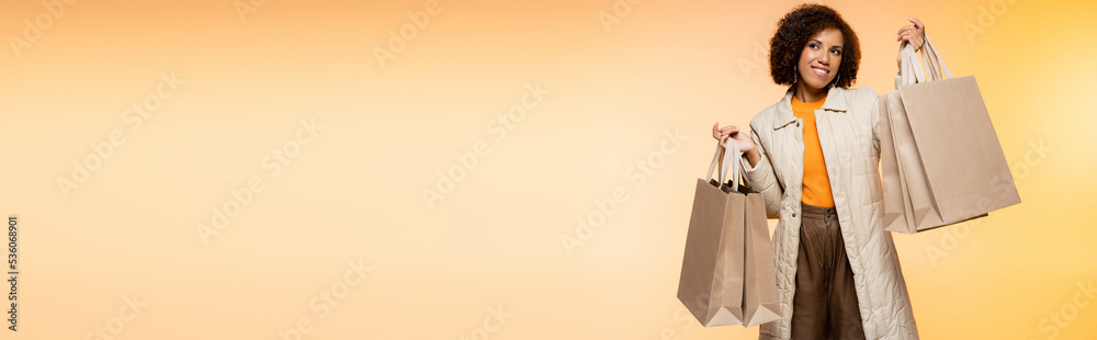 positive african american woman in coat holding black friday shopping bags on orange, banner. positive african american woman in coat holding black friday shopping bags on orange, banner.