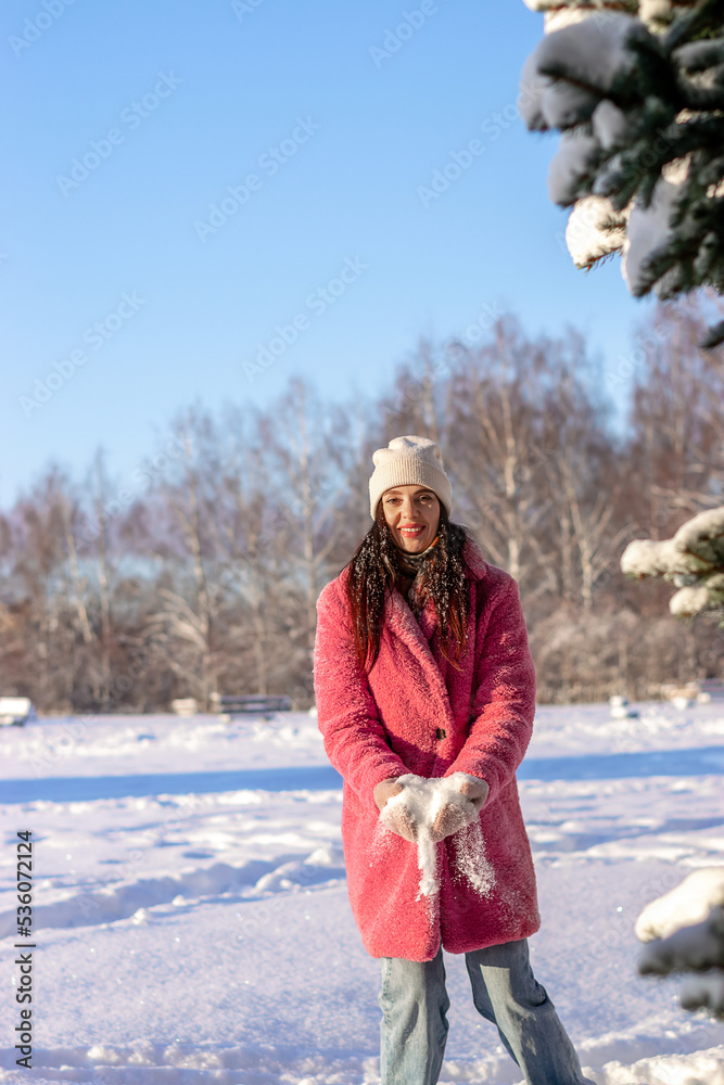 Fototapeta premium Young beautiful woman plays with snow in a winter park, throws it up and laughs.Winter concept.Selective focus,copy space.