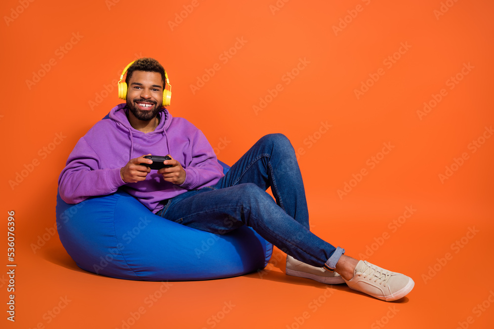 Full size portrait of cheerful handsome guy sit cozy bag enjoy playing games isolated on orange color background Full size portrait of cheerful handsome guy sit cozy bag enjoy playing games isolated on orange color background