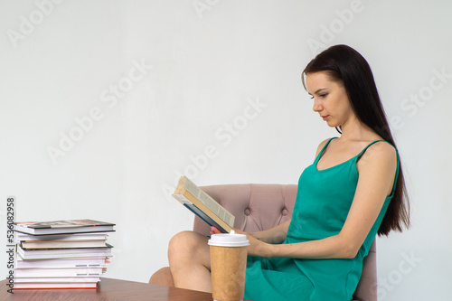 Молодая красивая девушка в зелёном платье читает книгу в кресле на белом фоне