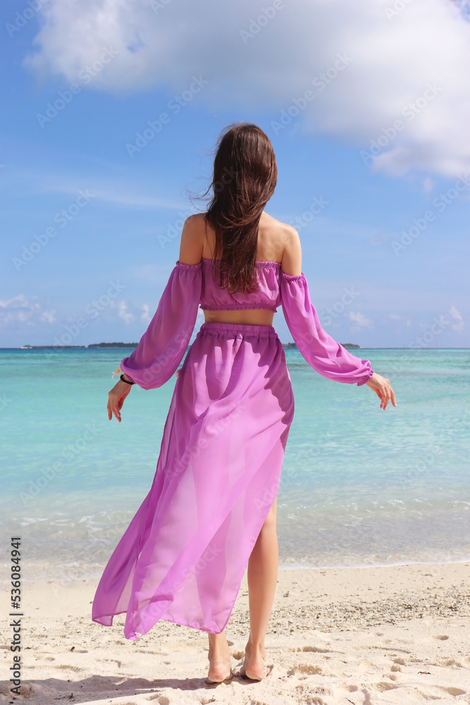 Luxury fashion. Elegant fashion model. Stylish female model in lilac beach cape, standing with her back to the beautiful turquoise ocean in the Maldives Luxury fashion. Elegant fashion model. Stylish female model in lilac beach cape, standing with her back to the beautiful turquoise ocean in the Maldives