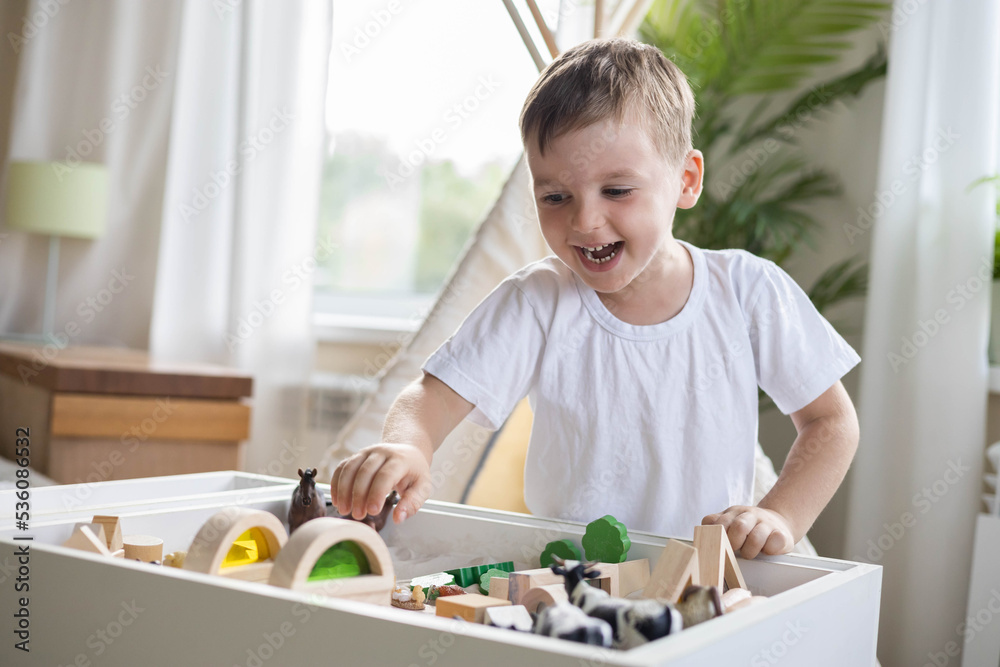 Smiling boy kid playing sensory box kinetic sand table with Montessori wooden materials Smiling boy kid playing sensory box kinetic sand table with Montessori wooden materials