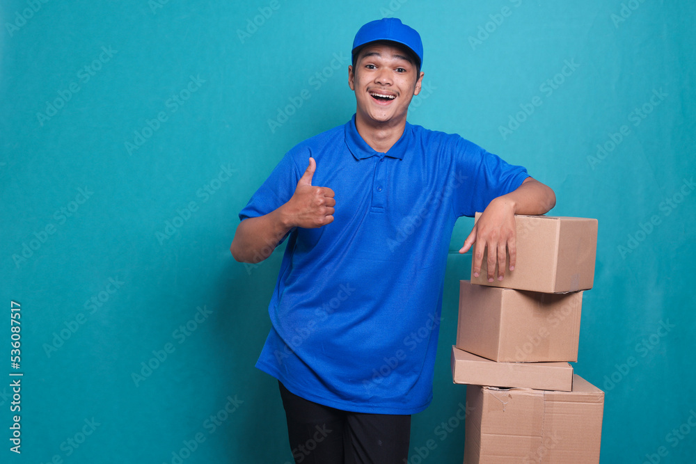 Delivery man in blue uniform work as dealer courier hold a stack of blank cardboard box and giving thumb up Delivery man in blue uniform work as dealer courier hold a stack of blank cardboard box and giving thumb up