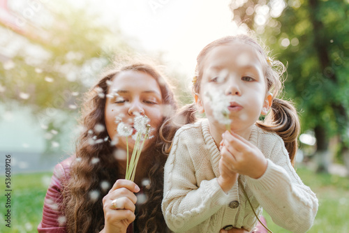 Young pretty women with small girl blowing dandelions in the park. Spend happy time together outside.