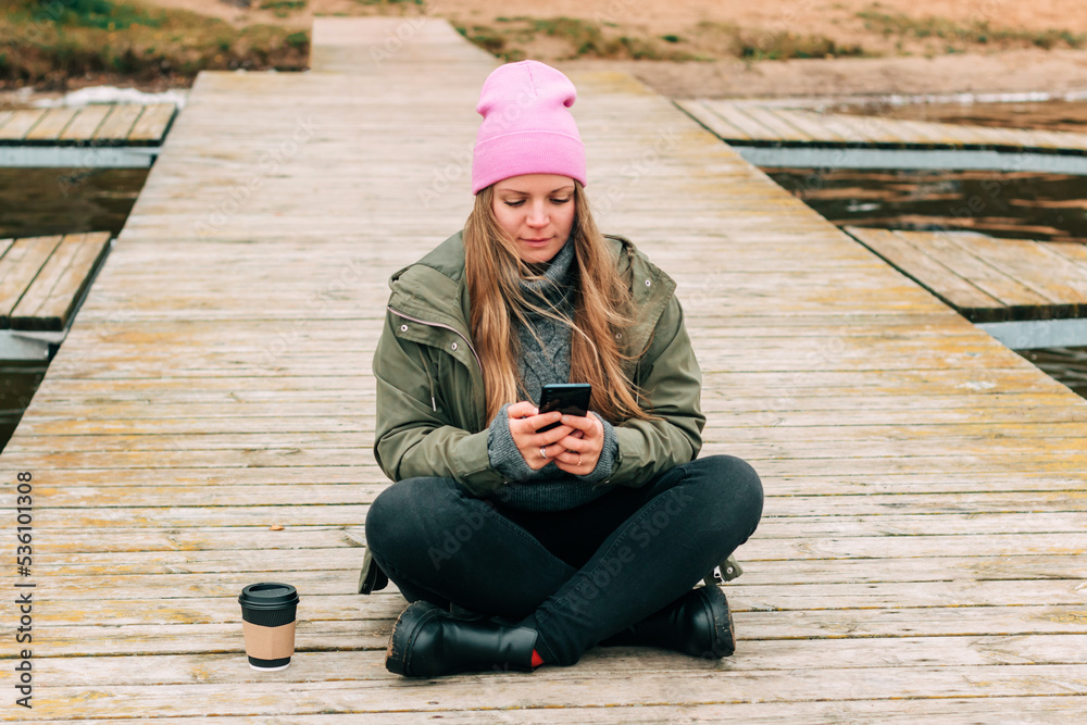 Woman in hat outdoor in the forest sitting on the bridge drinking a cup of hot coffee and using a mobile phone technology. Beautiful female relax and enjoy outdoor activity lifecycle in nature 