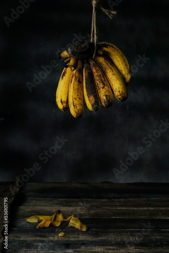 a bunch of ripe bananas hanging with a jute rope. dark background. wooden lining. there is a peeled banana skin on the wooden base.
