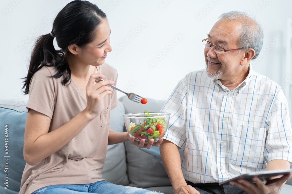 Happy Asian daughter making salad and feeding to senior father sitting on couch at home. Senior man smiling of tasty food from beloved girl. Family spending time together on weekend Happy Asian daughter making salad and feeding to senior father sitting on couch at home. Senior man smiling of tasty food from beloved girl. Family spending time together on weekend
