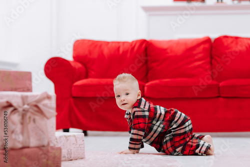 Adorable caucasian baby boy is crawling by the Christmas tree, many festive gift boxed and red sofa at home, cute infant in stylish pyjama. Xmas and New Year family celebrations concept. © Andriy Medvediuk