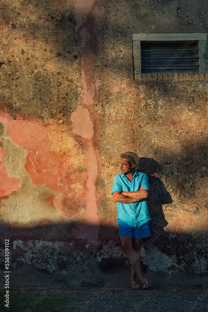 Obraz premium Blond man in blue shirt and swim shorts stands against weathered pink plaster wall in late afternoon sunlight.