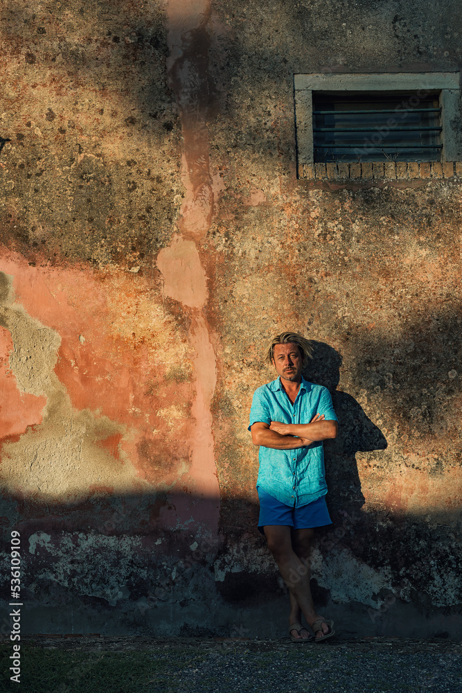Blond man in blue shirt and swim shorts stands against weathered pink plaster wall in late afternoon sunlight. Blond man in blue shirt and swim shorts stands against weathered pink plaster wall in late afternoon sunlight.