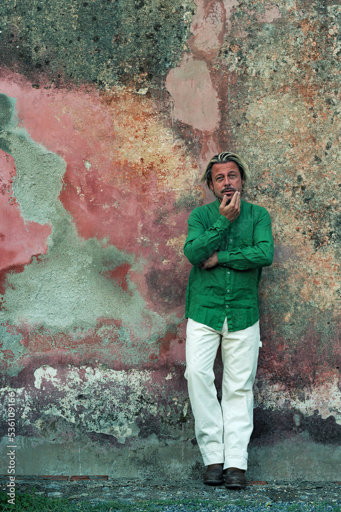 Obraz premium Blond tanned man in green shirt and white pants stands in front of weathered pink plaster wall.