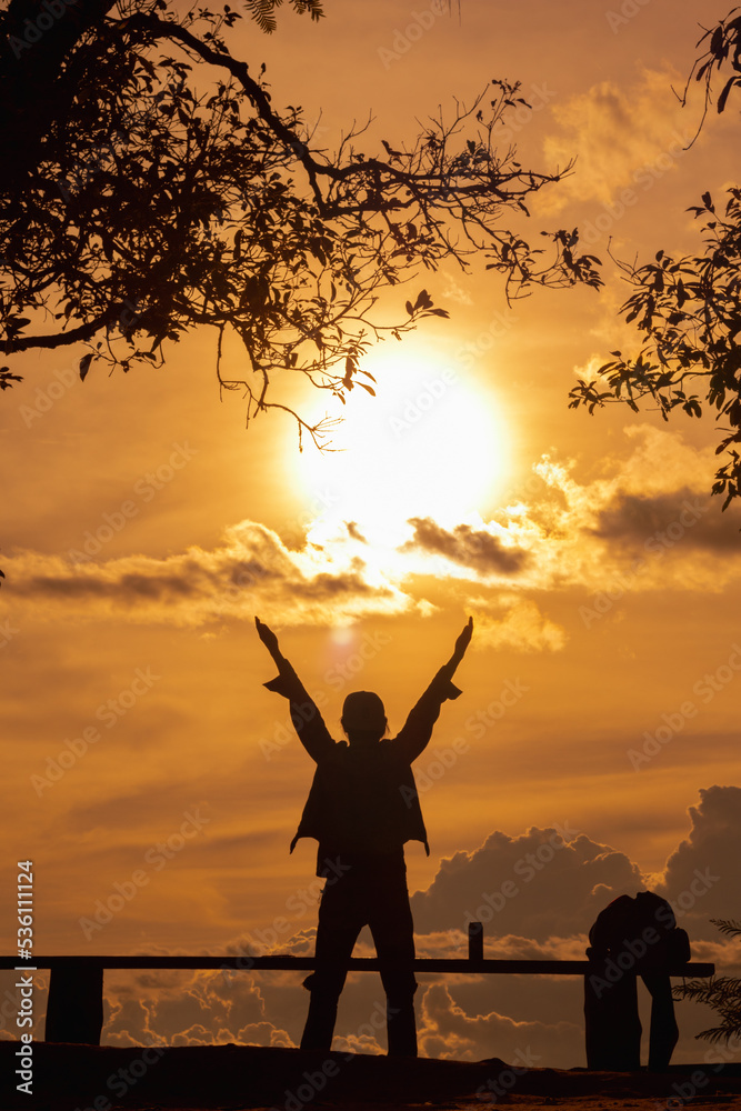 A silhouette of a young man with both hands raised above his head and praying to God with his faith and power on a blurry background of the beautiful view of the morning sun.