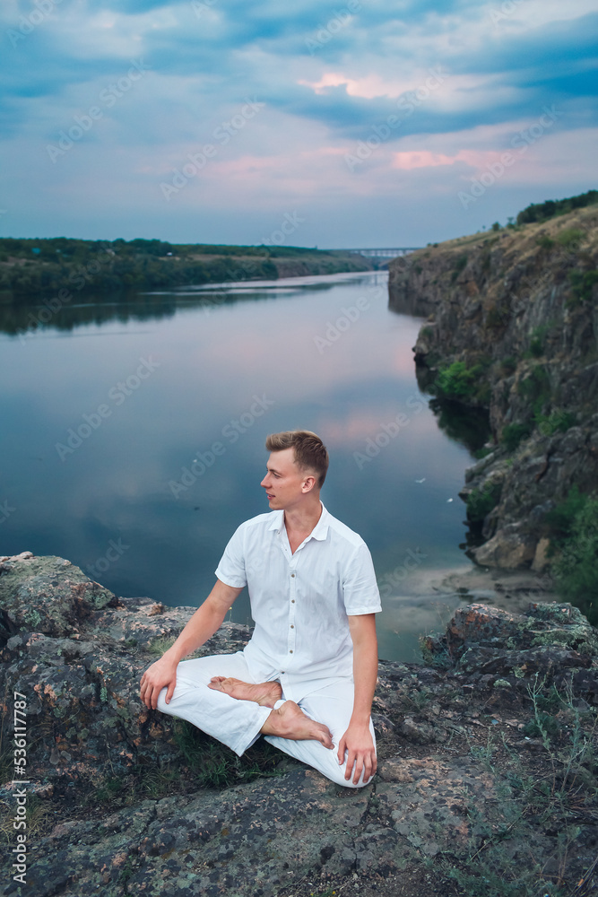 Fototapeta premium A handsome young guy is engaged in meditation sitting in a lotus position at dawn, morning meditation on the river bank.