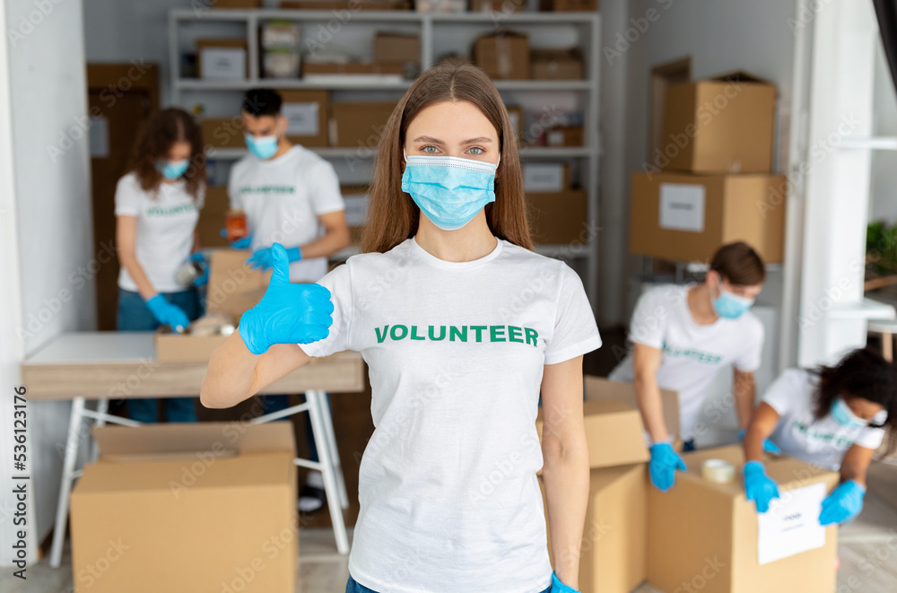 Female activist wearing medical mask, working at volunteer center, showing thumb up at camera Female activist wearing medical mask, working at volunteer center, showing thumb up at camera