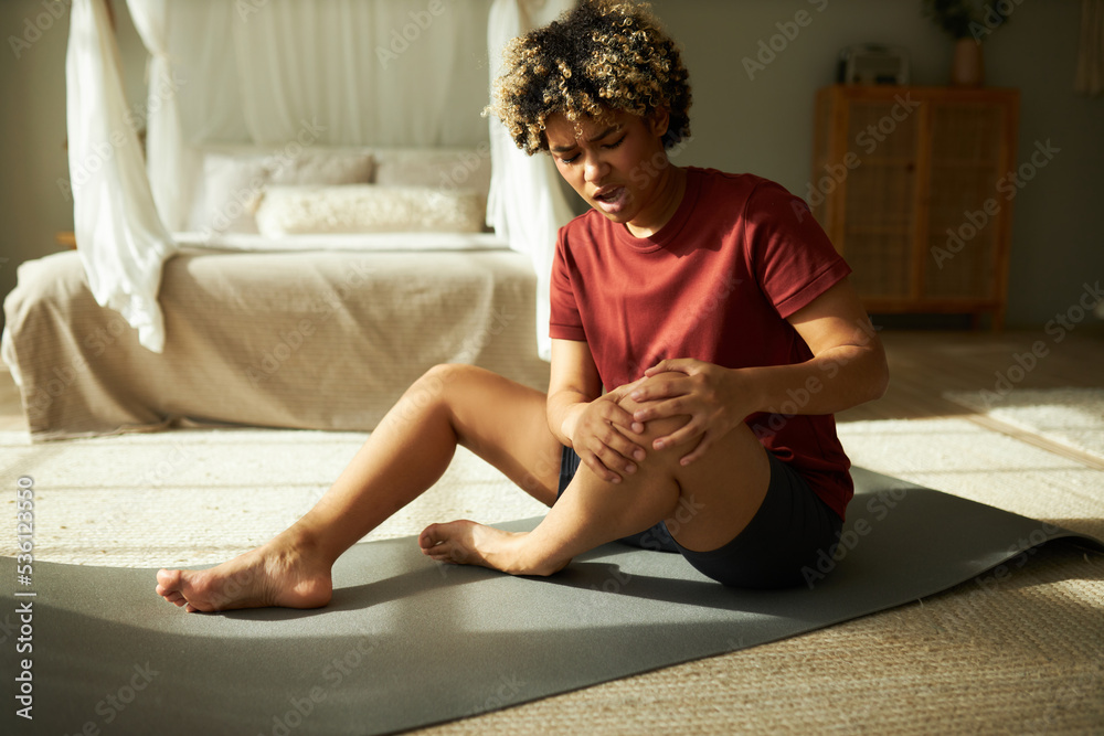 African american girl sitting on gray mat, grabbing hurt knee, unable to start yoga workout after getting sport injury, suffering from pain, female beginner doing wrong exercise without coach - obrazy, fototapety, plakaty 