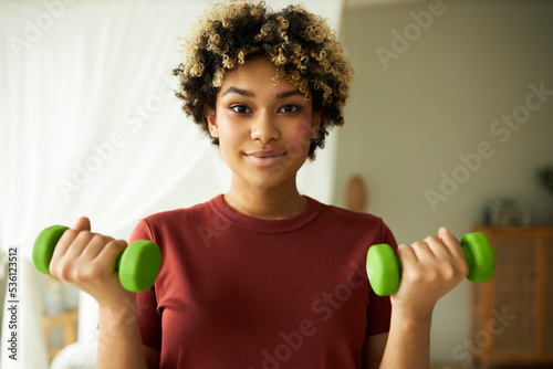 Closeup portrait of cute african american female with afro hair holding green dumbbells doing training workout at home, building muscles, wearing red t-shirt. Healthy lifestyle, fitness at home