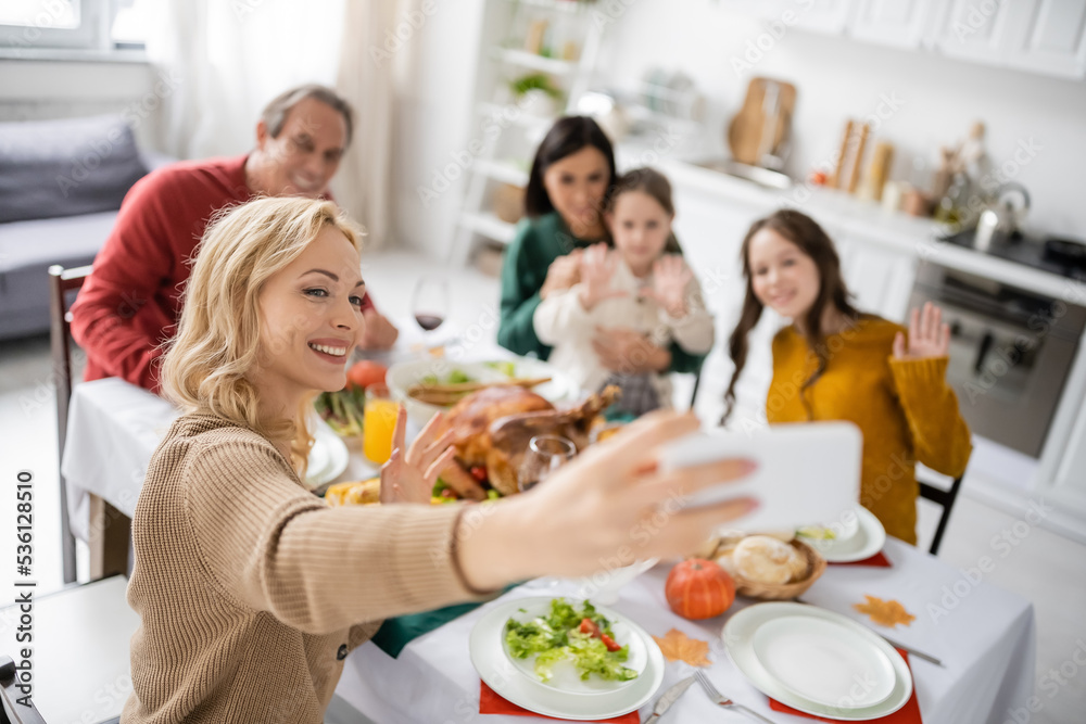 Smiling woman taking selfie with blurred family near thanksgiving dinner at home