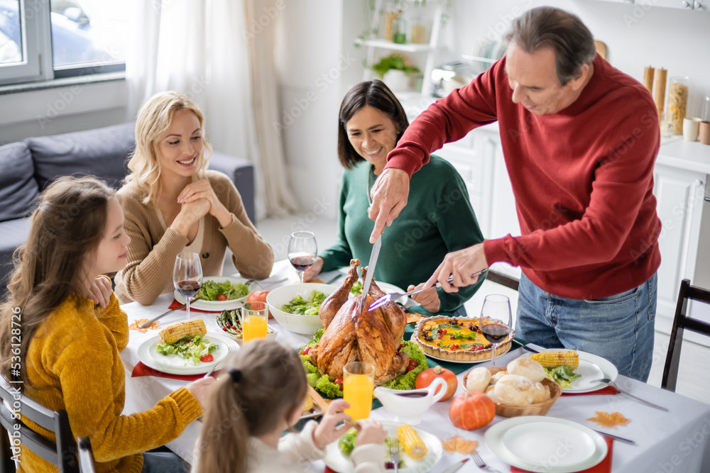 Man cutting turkey near smiling multiethnic family during thanksgiving dinner at home