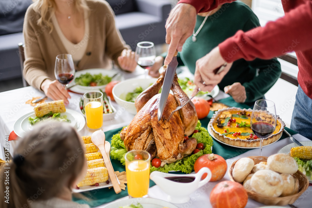 Man cutting turkey near blurred family and tasty thanksgiving dinner