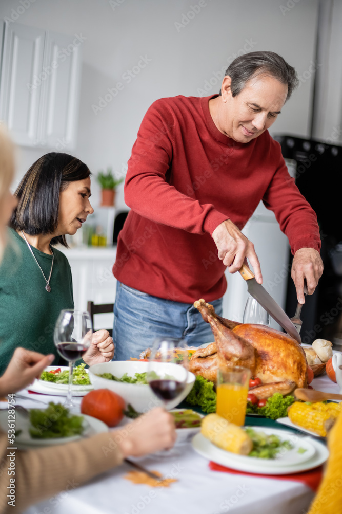 Senior man cutting turkey near interracial family during thanksgiving dinner at home