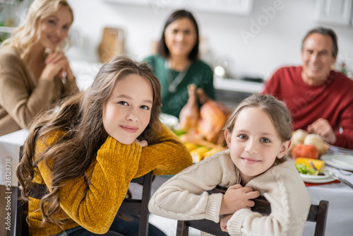 Smiling girls looking at camera near blurred family and thanksgiving dinner at home