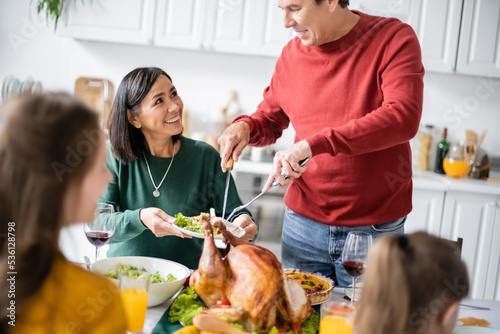 Senior man putting turkey on plate near interracial family celebrating thanksgiving at home