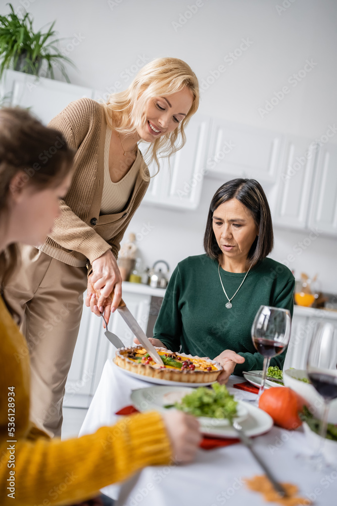 Smiling woman cutting delicious thanksgiving pie near multicultural mother and child at home
