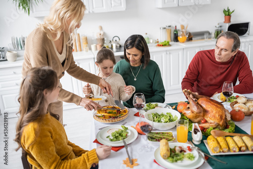 Multicultural family with kids looking at woman cutting thanksgiving pie during dinner at home