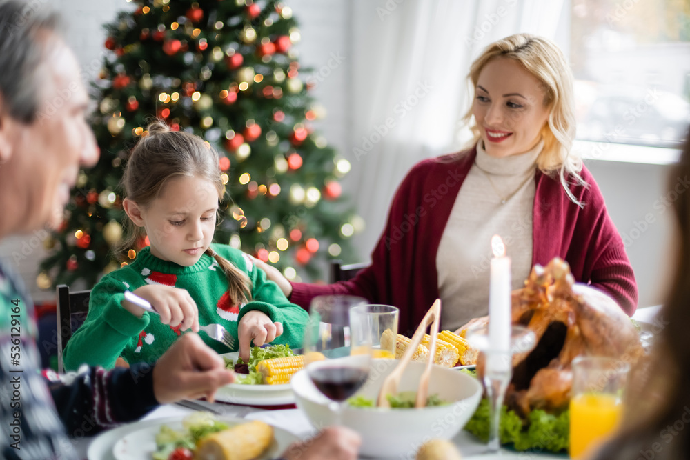smiling woman looking at daughter having festive family dinner near christmas tree