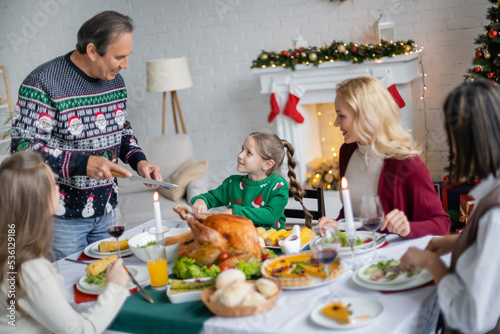 smiling girl looking at grandfather with knife while having christmas dinner with interracial family
