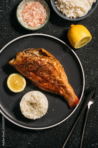 baked fish perch with lemon, boiled rice, spices nearby. fish on a black plate and dark background