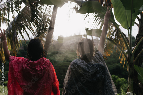 Happy girls in raincoats waving hands on nature background. Two female travelers expressing positive emotions during walk in tropical forest.