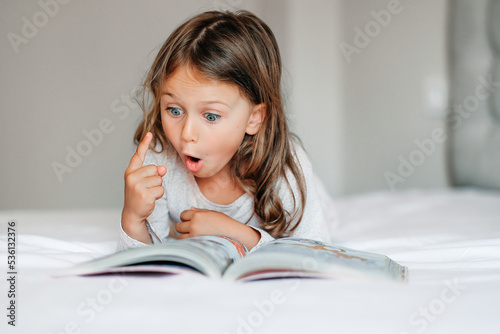 Emotional child girl read interesting book in bed. Small kid enjoy reading new facts pointing finger. Developing child fantasy and imagination. Imaginary world. Fairy tale. Bedtime reading photo