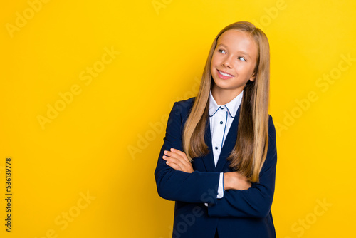 Portrait of positive schoolgirl with long hairstyle dressed blue jacket look empty space arms crossed isolated on yellow color background