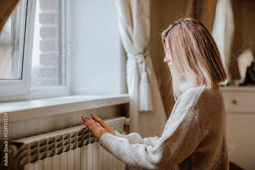 Young blond woman in long winter beige sweater is posing near the radiator.