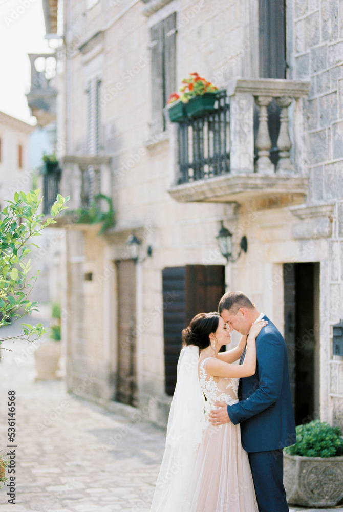 Groom hugs bride in a white dress near a stone building with balconies