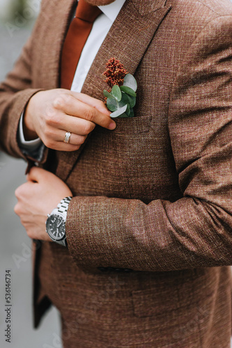 Close-up portrait of a groom in a brown, expensive, stylish suit with a boutonniere, tie, wristwatch and a gold ring on his finger. wedding photography.
