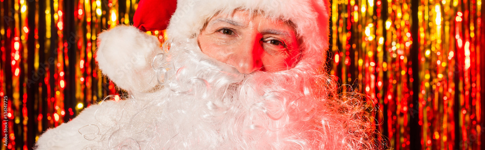 Portrait of santa claus in red hat looking at camera near blurred tinsel, banner.