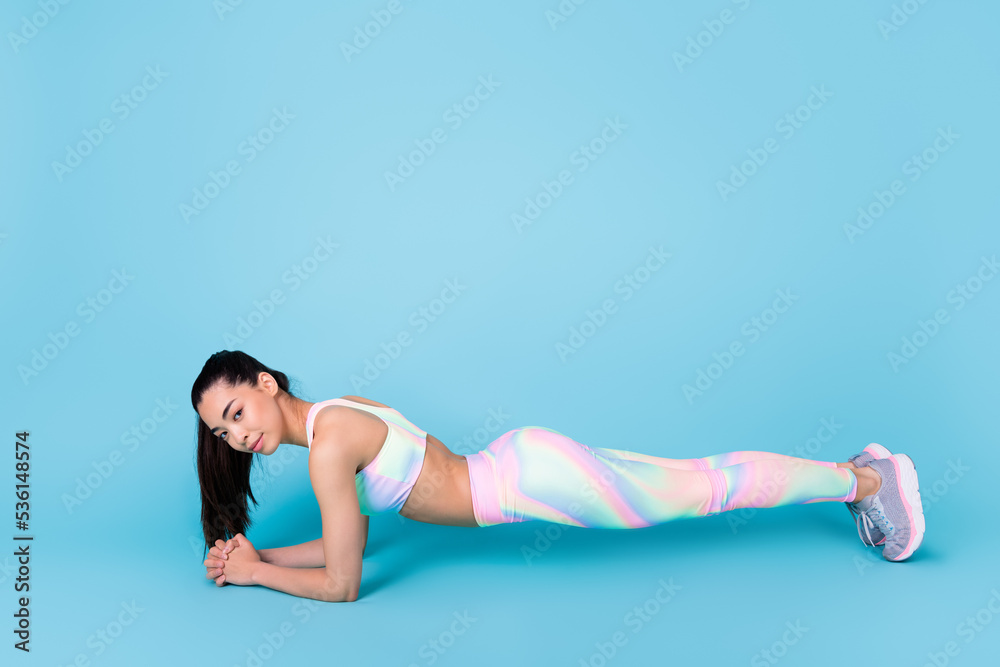 Full length photo of positive pretty lady wear sport suit standing plank empty space isolated blue color background Full length photo of positive pretty lady wear sport suit standing plank empty space isolated blue color background