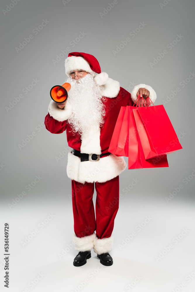 Santa claus in costume and eyeglasses holding loudspeaker and shopping bags on grey background. Santa claus in costume and eyeglasses holding loudspeaker and shopping bags on grey background.
