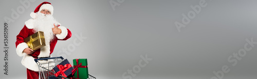 Father christmas showing thumb up near shopping cart with presents isolated on grey, banner.
