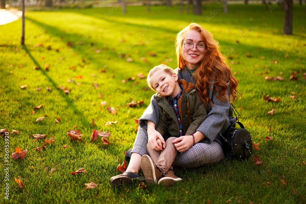 Smiling redhead woman and little boy sitting on grass lawn in city park on warm autumn day. Young mom hugs her son, they have fun and look at camera on sunny fall day. Foliage on green lawn. Smiling redhead woman and little boy sitting on grass lawn in city park on warm autumn day. Young mom hugs her son, they have fun and look at camera on sunny fall day. Foliage on green lawn.
