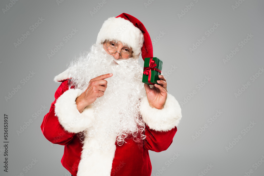 Santa claus in eyeglasses pointing at small gift box isolated on grey.
