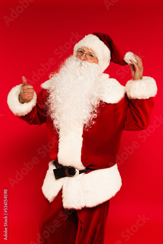 Santa claus touching hat and showing like gesture on red background.