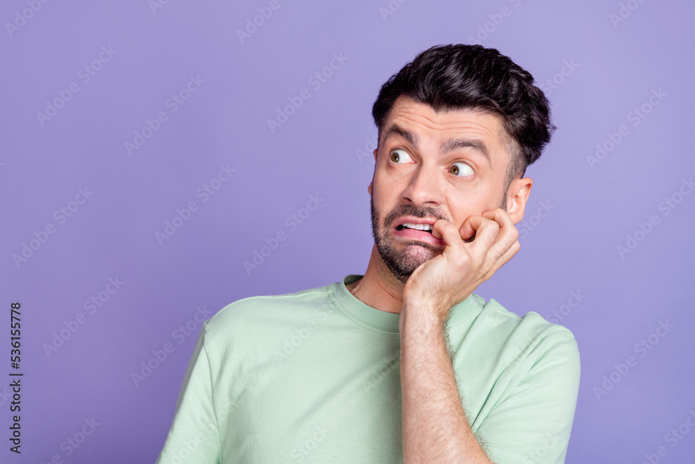 Portrait of terrified horrified man with brunet hairdo dressed t-shirt biting fingers look empty space isolated on purple color background