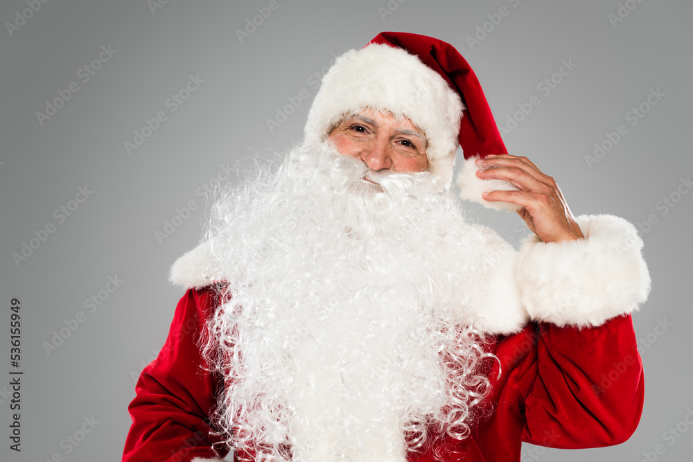 Santa claus in costume touching hat and looking at camera isolated on grey.