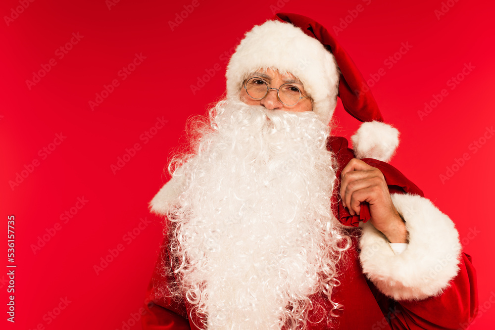 Bearded santa claus holding sack and looking at camera isolated on red.