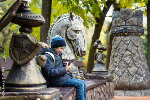 a boy sits on a bench among sculptures in the form of chess pieces in the park, plays online chess on his phone. in Presnensky park