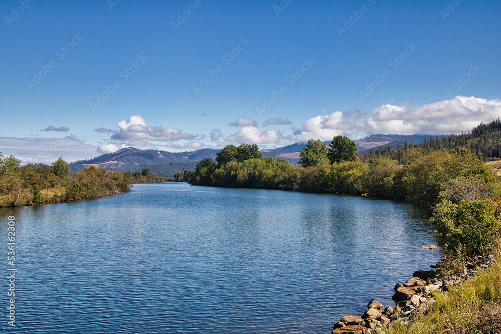 On a sunny Autumn day in Northwestern Idaho along the Trail of the Coeur d'Alenes, the partly cloudy blue sky is reflected in the surface of a peaceful river with mountains in the distance.