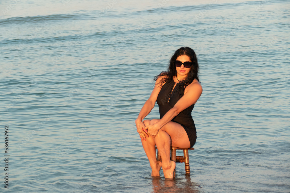 The girl is sitting on a stool in the sea. Brunette in a black swimsuit on the beach.